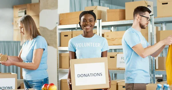 Retrato de una hermosa joven afroamericana alegre sostenida en caja de donaciones de manos, mirando a la cámara con sonrisa en la cara. Actividad benéfica Voluntarios empacando comida y ropa Concepto de voluntariado —  Fotos de Stock