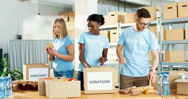 Mujeres y hombres voluntarios afroamericanos y caucásicos ocupados preparando cajas de donaciones, empacando alimentos y comestibles. Actividad caritativa. Voluntariado y donación. Ayuda social concepto de trabajo —  Fotos de Stock