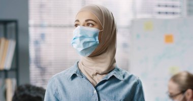 Tıp maskeli genç Arap kadın çalışanın yakın plan portresi. Ofiste durmuş, keyifli bir şekilde kameraya bakıyor. Kabinede çalışan Arap kadın işçi, koronavirüs salgını konsepti