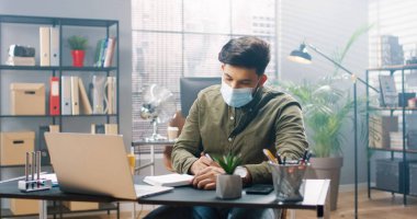 Tıbbi maskeli genç bir adam ofiste tek başına oturur ve dizüstü bilgisayarındaki bilgileri incelerken defterine notlar alır..