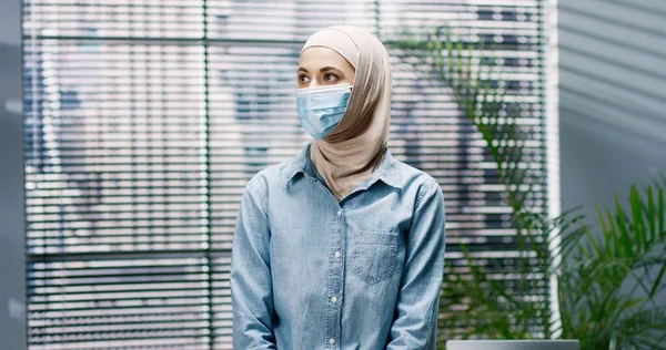 Retrato de la joven árabe hermosa mujer en máscara médica de pie en el interior de la oficina mirando a la cámara en el trabajo. Empleada en el gabinete en cuarentena, cierre del coronavirus, concepto de ocupación —  Fotos de Stock