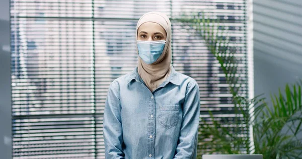 Retrato de la joven árabe hermosa mujer en máscara médica de pie en el interior de la oficina mirando a la cámara en el trabajo. Empleada en el gabinete en cuarentena, cierre del coronavirus, concepto de ocupación —  Fotos de Stock