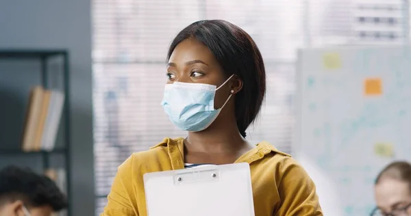 Закріпіть портрет молодої афроамериканської працівниці в медичній масці, що стоїть при владі, дивлячись на камеру і посміхаючись. Радісна жінка менеджер в гарному настрої, коронавірська концепція — стокове фото