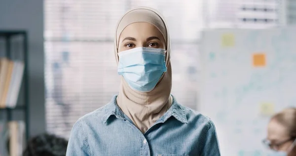 Primer plano retrato de joven árabe hermosa empleada en máscara médica de pie en la oficina en el trabajo, mirando a la cámara de buen humor. Trabajadora árabe en el gabinete, concepto pandémico de coronavirus — Foto de Stock