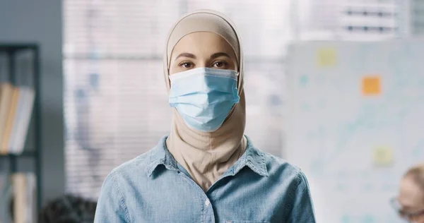 Primer plano retrato de la joven hermosa mujer en hijab y máscara médica protectora de pie en la oficina y mirando a la cámara. — Foto de Stock