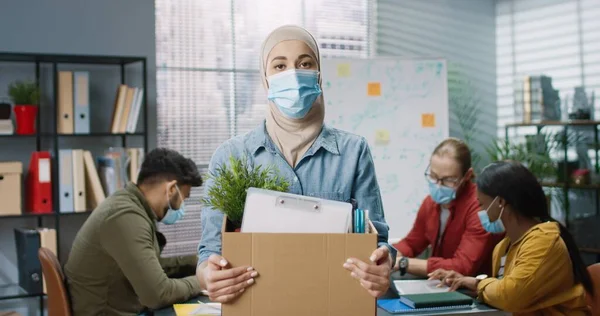 Retrato de una mujer árabe hermosa trabajadora con máscara médica parada en la oficina sosteniendo una caja de cartón con sus cosas acaba de ser despedida del trabajo. Trabajadores de raza mixta que trabajan detrás en la oficina, concepto de empleo — Foto de Stock