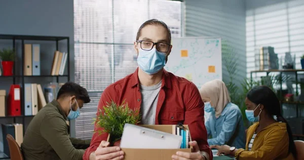 Retrato de joven caucásico guapo trabajador masculino en máscara médica de pie en la oficina sosteniendo caja de cartón con sus cosas acaba de ser despedido del trabajo Los trabajadores de raza mixta que trabajan detrás en la oficina, desempleo — Foto de Stock