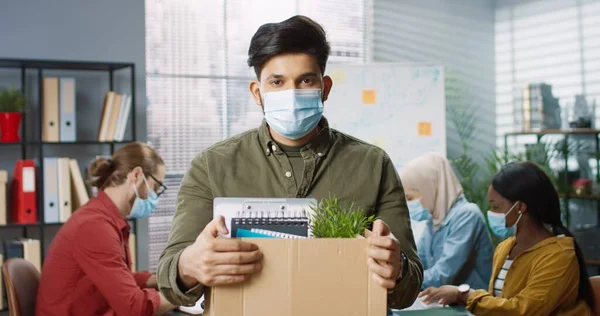 의료용 마스크를 입은 젊고 잘생긴 힌두교 남성 이 자신의 물품이든 상자를 들고 캐 비넷 안에 서 있는 모습 이 직장에서 해고되었다. 혼합 인종 노동자 사무실, 고용 개념 뒤에서 일하는 — 스톡 사진