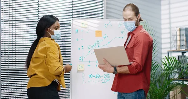 Dos colegas multinacionales están trabajando en una tarea conjunta en la pizarra blanca con máscaras protectoras, creando infografías usando un cuaderno. — Foto de Stock