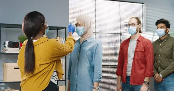 Ritratto di donna afroamericana in maschera medica in piedi in ufficio a controllare la salute di diversi lavoratori di razza mista che misurano la temperatura utilizzando il termometro a infrarossi durante la pandemia di coronavirus — Foto Stock