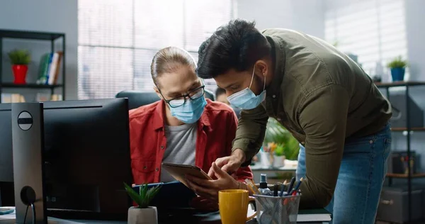 의료용 마스크를 쓰고 있는 힌두교 남성 이 백인 청년 동료에게 와서 직장에서 무엇인가를 논의하고 있는 태블릿 장치에 무엇인가를보여 주고 있다. 사무실 컨셉, 회사 일 — 스톡 사진