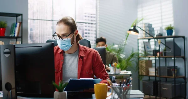 의료용 마스크를 쓰고 책상에 앉아 컴퓨터 타이핑을 하고 있는 백인잘생긴 남성 노동자의 모습. 직장에서 사업 상의 문제에 대해 상사에게 이야기하는 것을 보여 주는 여성 동료. 사무실 컨셉 — 스톡 사진