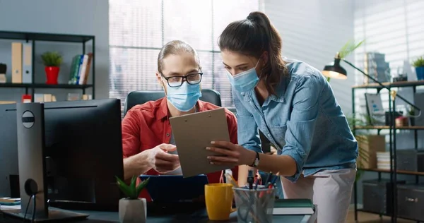 Dos colegas en máscaras médicas protectoras consultan mientras resuelven el problema durante el proceso de trabajo mientras están sentados en la oficina. — Foto de Stock