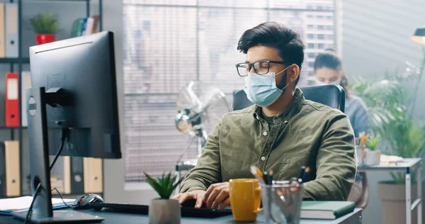 Joven hombre exitoso en gafas y máscara protectora se sienta en la oficina en su escritorio y trabaja intensamente en la computadora. — Foto de Stock