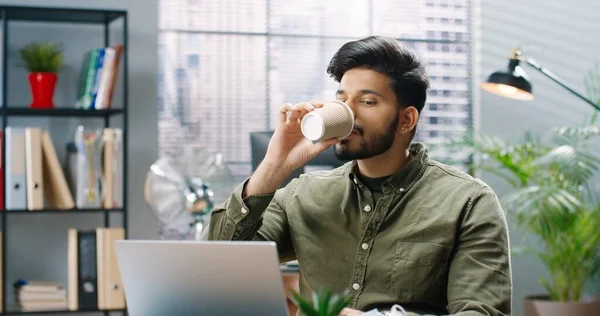 Ung stilig man sitter på kontoret vid bordet dricka kaffe sätter på skyddande medicinsk mask och fortsätter att skriva på laptop. — Stockfoto