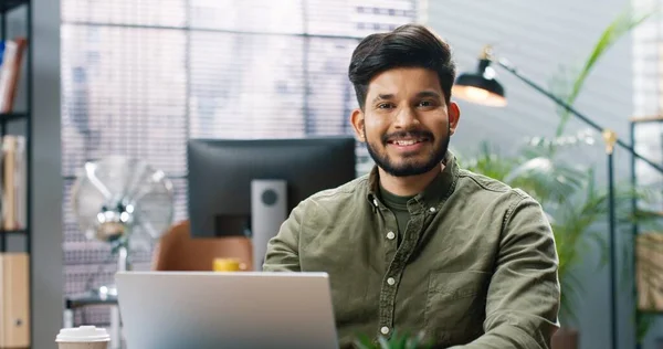 Närbild porträtt av glad snygg ung hinduisk manlig arbetare sitter vid skrivbordet i moderna skåp och knacka på laptop surfa online på kontoret ta bort medicinsk mask tittar på kameran med leende på ansiktet — Stockfoto