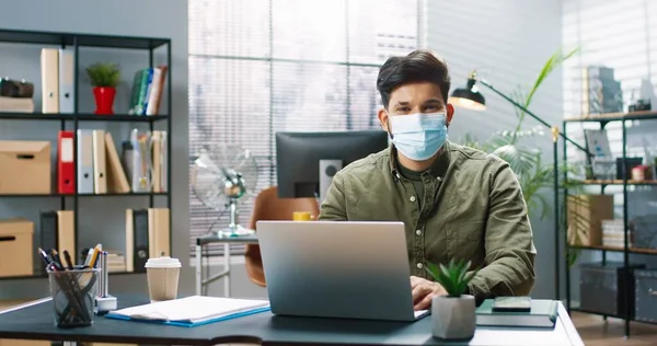 Портрет молодого красивого мужчины в защитной медицинской маске, сидящего за рабочим столом и работающего на ноутбуке, улыбающегося в камеру. — стоковое фото