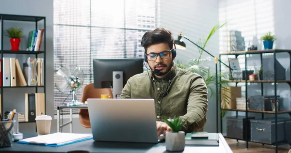 Porträtt av stilig hinduisk ung skäggig man talar i headset och skriva på laptop medan du sitter i skåp på arbetsplatsen i moderna skåp. Kundservicearbetare, affärsidé — Stockfoto