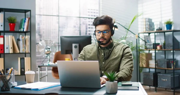 Ung stilig hinduisk man i glasögon och hörlurar med mikrofon sitter på kontoret ensam och arbetar på laptop. — Stockfoto