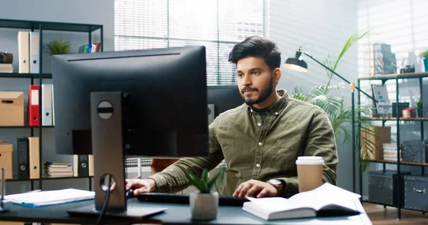 Porträtt av ung hinduisk stilig skäggig affärsman sms:ar på datorn söker internet på nätet medan du sitter på arbetsplatsen på moderna kontor. Framgångsrikt företag, kontorsarbete, yrkeskoncept — Stockfoto