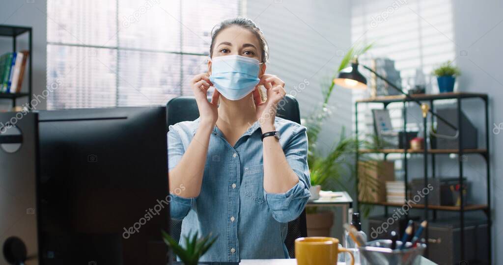 Portrait of young beautiful girl in blue shirt sitting in office at table and takes off protective medical mask smiling looking at camera.