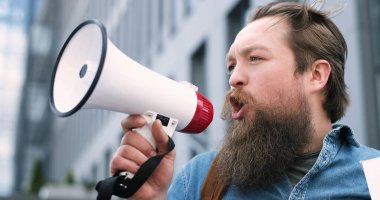 Close up of Caucasian young man with beard speaking and screaming slogans and mottos in megaphone outdoor. Single political protest. Male activist talking in speaker. clipart