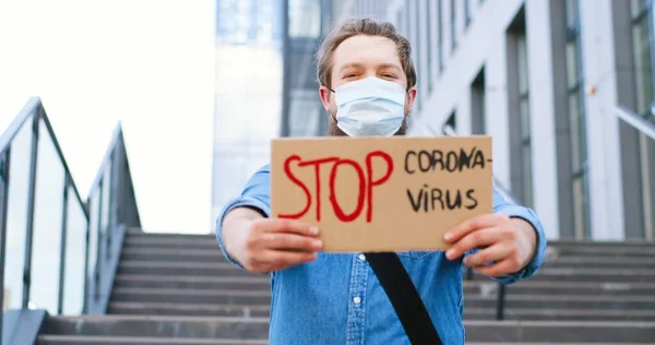 Portré fotó fehér fiatalemberről orvosi maszkban, kint áll a lépcsőnél, és táblát mutat szavakkal: Stop Coronavirus. Férfi bemutató tábla tiltakozó ellen világjárvány. — Stock Fotó