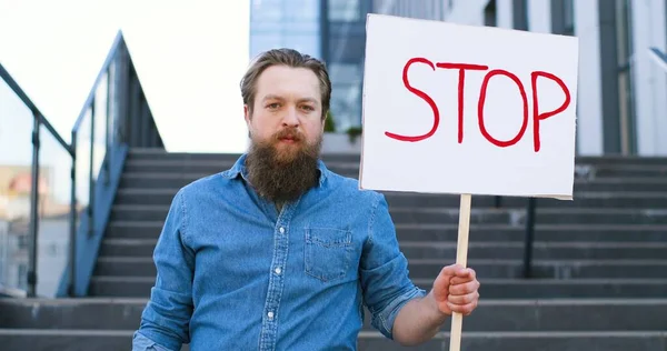Portret van een blanke mannelijke activist met een baard die een poster vasthoudt Stop bij een politieke of ecologische eenzame demonstratie. Enkel protest buiten. Activisme concept. Een man die alleen protesteert. — Stockfoto
