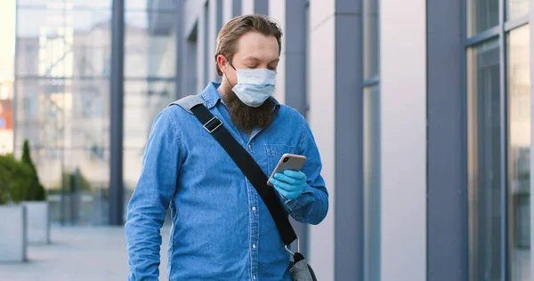 Hombre caucásico con barba y máscara médica caminando por la calle y enviando mensajes de texto en el teléfono móvil. Peatón masculino en protección respiratoria paseando al aire libre y tocando o desplazándose en el teléfono inteligente . — Foto de Stock