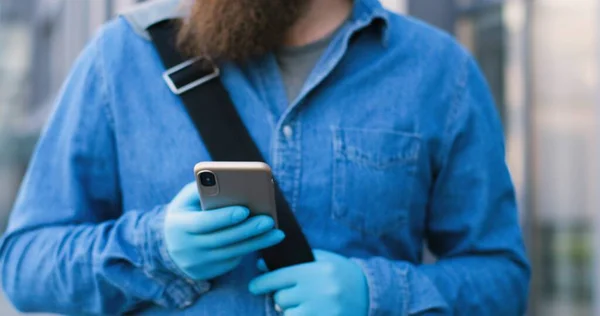 Close up of Caucasian man with beard and in medical mask texting message on mobile phone at street. Male pedestrian in respiratory protection tapping and scrolling on smartphone outdoors.