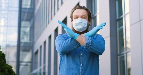 Retrato de un joven caucásico con máscara médica y guantes de pie en la calle y sin gesto prohibido con las manos. Varón cruzando brazos como parada, negativa o prohibición. Concepto pandémico . —  Fotos de Stock