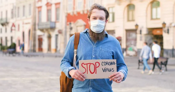 Retrato de un activista caucásico con barba y máscara médica sosteniendo un afiche de Stop Coronavirus en una manifestación solitaria en la ciudad. Una sola protesta afuera. Concepto de activismo. Hombre protestando solo . —  Fotos de Stock