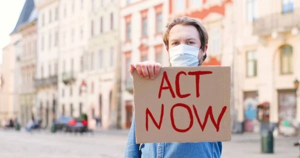 Retrato del cartel de cartón con palabras Act Now en manos de un joven caucásico con máscara médica. Protesta sola al aire libre en la plaza del pueblo. Activista sanitario masculino mostrando mesa . —  Fotos de Stock