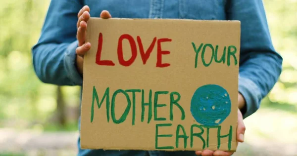 Feche o cartaz do cartão com palavras Ame sua Mãe Terra nas mãos do homem caucasiano. Macho eco ativista em pé na floresta ou parque no dia ensolarado com protesto único . — Fotografia de Stock