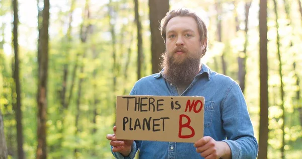 Portretfoto van blanke jonge mannelijke eco acivist die in het groene bos staat en met woorden naar de camera poster.Er is geen planeet B. Er is geen planeet. Knappe man die protesteert voor een schone en veilige omgeving. wazig. — Stockfoto