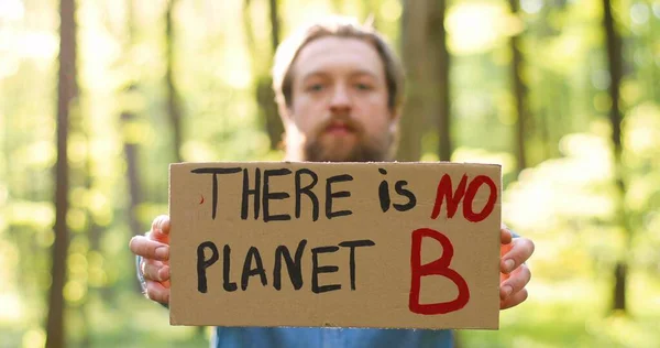 Retrato filmado de branco jovem eco acivista masculino em pé na floresta verde e subindo para o cartaz da câmera com palavras Não há planeta B Homem bonito protestando por um ambiente limpo e seguro. Desfocado . — Fotografia de Stock