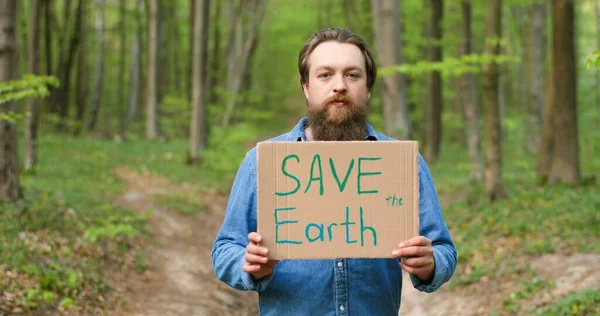 카프카스계 젊은 남성에 코 산악인 이 녹색 숲에 서서 지구를 구하라 는 문구가 적힌 포스터를 들고 있는 사진입니다. 깨끗하고 안전 한 환경을 위해 항의하는 친절 한 사람. — 스톡 사진