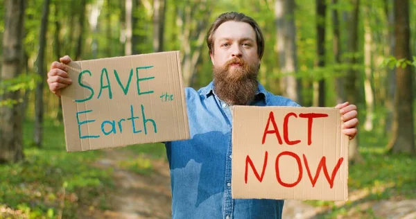 Porträtt av kaukasiska unga manliga eko acivist står i grön skog och stigande affischer med ord och stavning Rädda jorden och agera nu. Snygg man protesterar för ren och säker miljö — Stockfoto