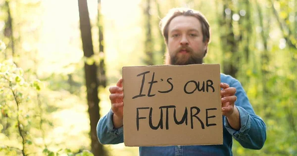 Portretfoto van een blanke jonge mannelijke eco-acivist die in het groene bos staat en een poster vasthoudt met woorden Its our Future. Knappe man protesteert voor schone en veilige omgeving. — Stockfoto