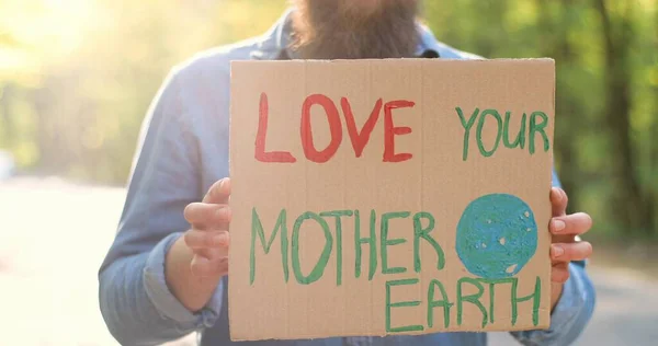 Zamknij kartonowy plakat w promieniach słońca słowami "Kochaj swoją Matkę Ziemię w rękach białego mężczyzny". Mężczyzna eko aktywista stojący w lesie lub parku w słoneczny dzień z pojedynczym protestem. Jasne światło słoneczne. — Zdjęcie stockowe