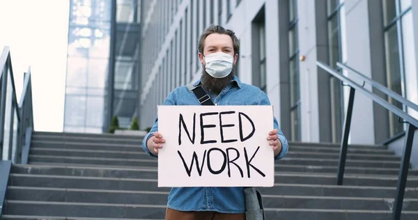 Joven activista masculino caucásico en mas medico parado en la calle de la ciudad en grandes escaleras con cartel Need Work. Tiempos pademicos duros. Concepto de protesta por desempleo . —  Fotos de Stock