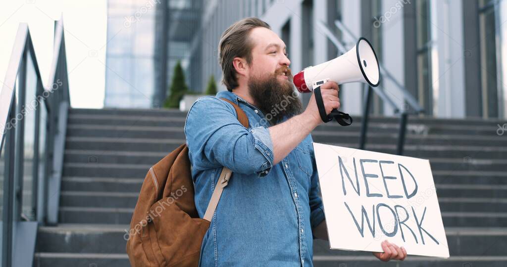 Portrait of Caucasian male activist with beard screaming in megaphone speaker. Man holding poster Need work at political lonely demonstration. Single protest after lockdown outdoors. Activism concept.