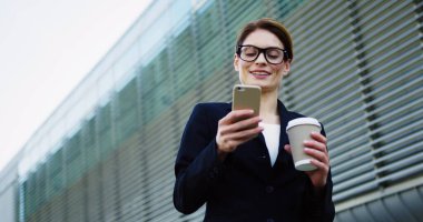 Beyaz, siyah ceketli, gözlüklü, akıllı telefonu bantlayan ve sabah kahvesini içen güzel bir iş kadını. Arkasında büyük bir ofis binası var. Açık havada.