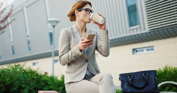 Beyaz tenli, gözlüklü bir iş kadını bankta kahve içerken akıllı telefonuyla kayıyor ve bantlıyor. Açık havada. — Stok fotoğraf