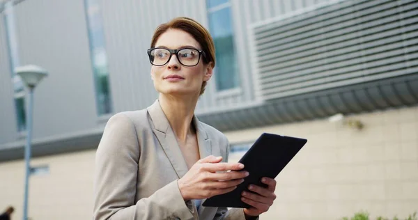 Close up of the attractive caucasian business lady in glasses scrolling and taping on the tablet computer while sitting on the bench, then looking at the side. Outside — Stock Photo, Image