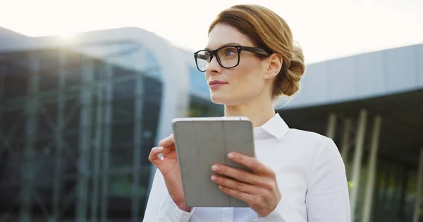 Портрет белой женщины в очках и белой рубашке, наклеенной на планшетное устройство и смотрящей на боковую сторону возле стеклянного офисного здания. На открытом воздухе — стоковое фото