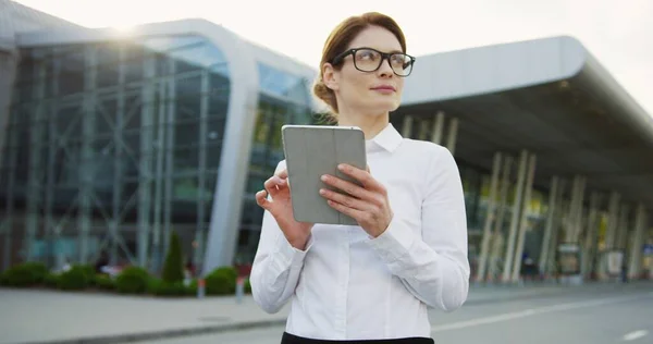 Portrait shot of the Caucasian smiled attractive woman in glasses and white shirt scrolling on the tablet computer and looking at the side near glass office building. Outside — Stock Photo, Image
