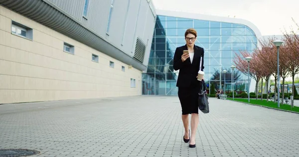 Vážná sebevědomá obchodní dáma v brýlích a černém obleku, jak jde do práce, nosí dokumenty a kávu, telefonuje a pak ze složky vytahuje papíry. Venku — Stock fotografie
