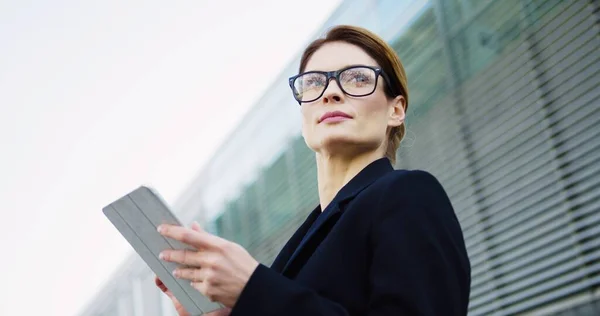 Close up of the Caucasian good looking woman in the glasses taping on the tablet device, then looking at the side near big building. Outdoor — Stock Photo, Image