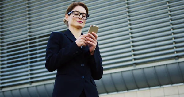 Portrait shot of the Caucasian pretty business lady in the glasses and black jacket taping on the smartphone, smiling and looking at the side near big office center. Outdoor — Stock Photo, Image